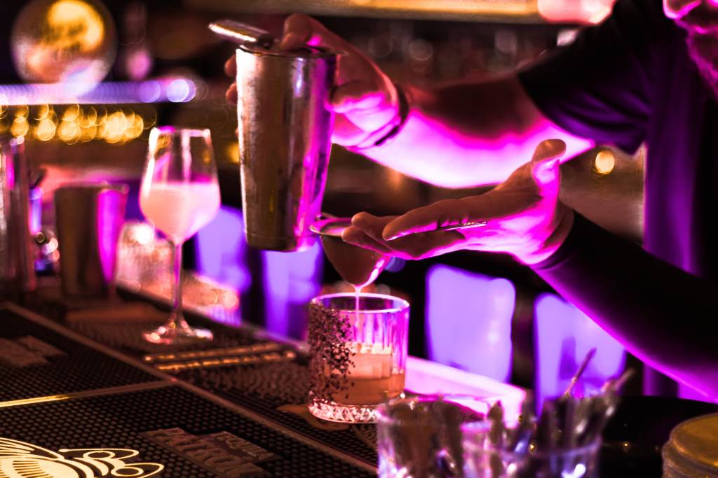 A close-up of a bartender pouring a drink, illuminated by colorful lights.