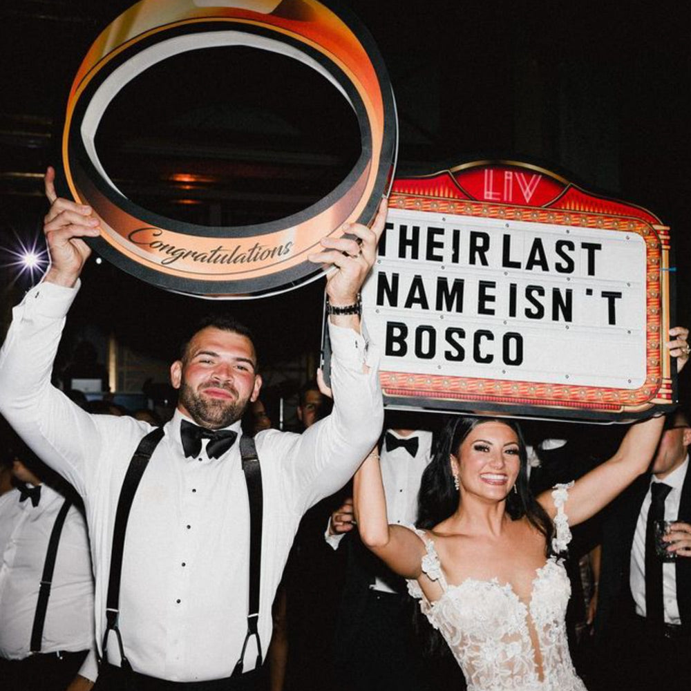 Bride and groom holding a Wedding Party Sign with a custom message and an engagement ring neon sign.