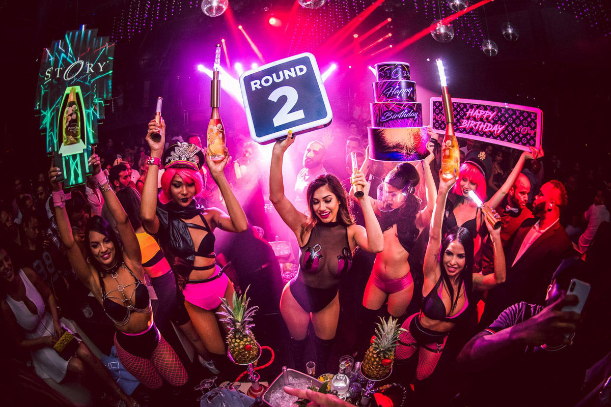 Crowd dancing at a club with pink neon lights and bottle service signs.